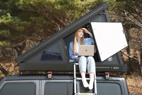 Black rooftop tent from which a woman with laptop looks out