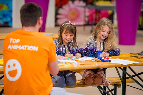 2 young girls in glitter jackets sign at a table in front of them sits someone from the entertainment team